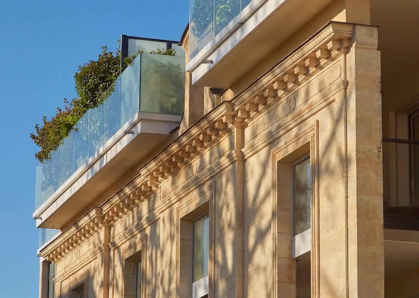 Balcon en verre moderne sur façade en pierre avec détails architecturaux classiques, sous un ciel bl.